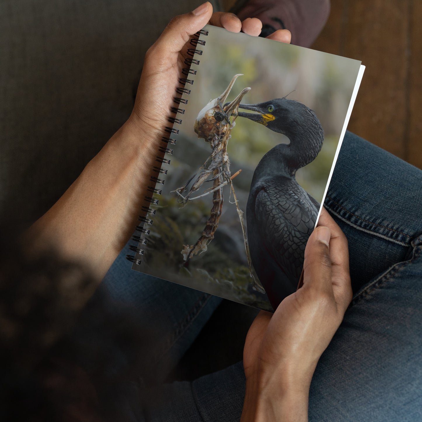 Spiral notebook with on the cover a picture of a shag holding another shag's skeleton in his beak. The notebook is being held by someone sitting on a couch in jeans.