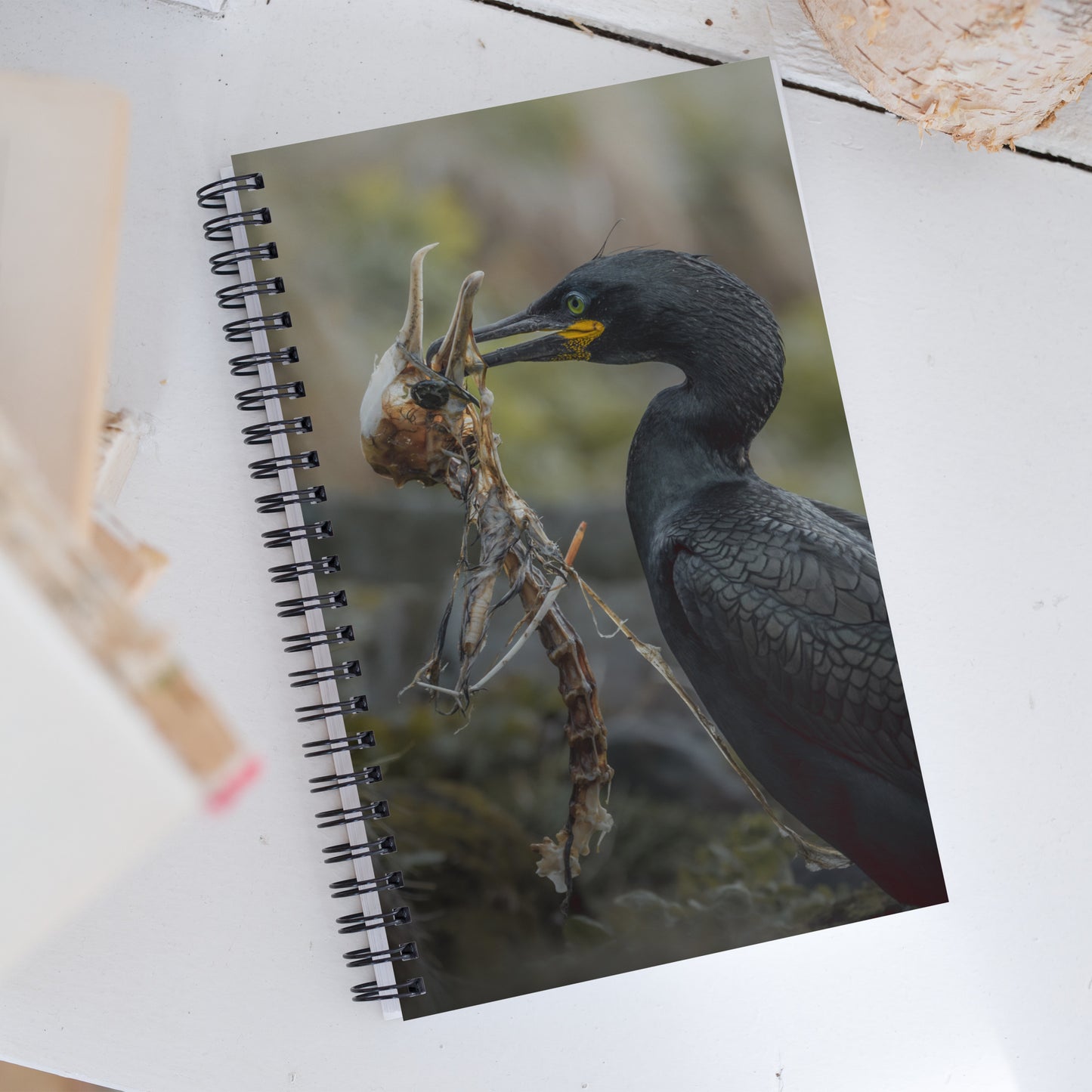 Spiral notebook with on the cover a picture of a shag holding another shag's skeleton in his beak. The notebook is laying on a white table.