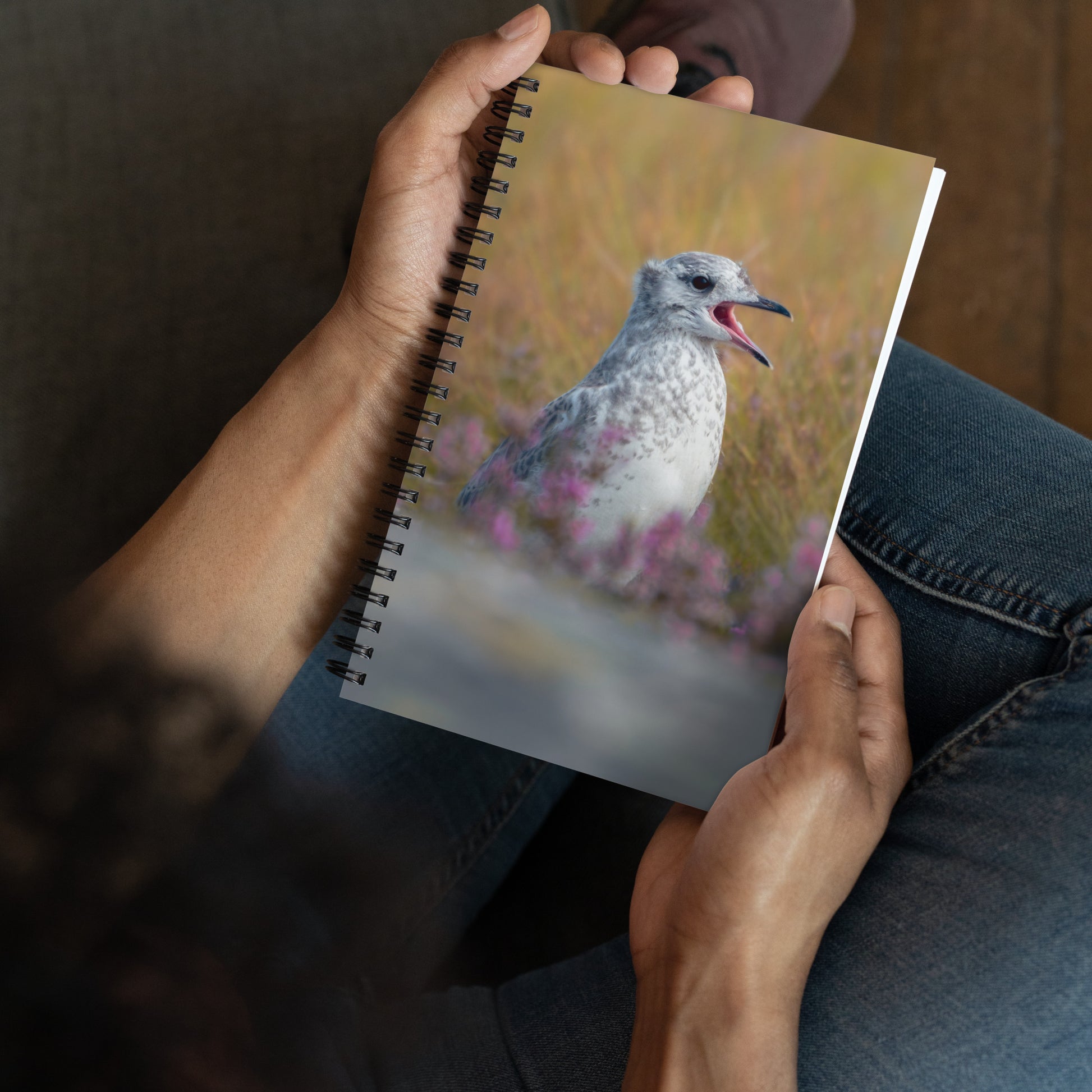 Spiral notebook with on the cover a picture of a young common gull surrounded by green grass and pink flowers.