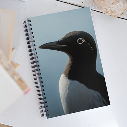 Spiral notebook with on front a portrait of a bridled common murre: a medium sized seabird with a white belly, black back ajnd head, medium-long and sharp black beak, and a white circle and line (resembling glasses) around the eyes.